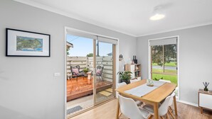 A modern interior with a wooden dining table, white chairs, a bookshelf, and a sliding glass door leading to an outdoor patio with a wooden deck and chairs. - Parrwang Rest - Renovated cosy unit in a Serene Location. (Portarlington)