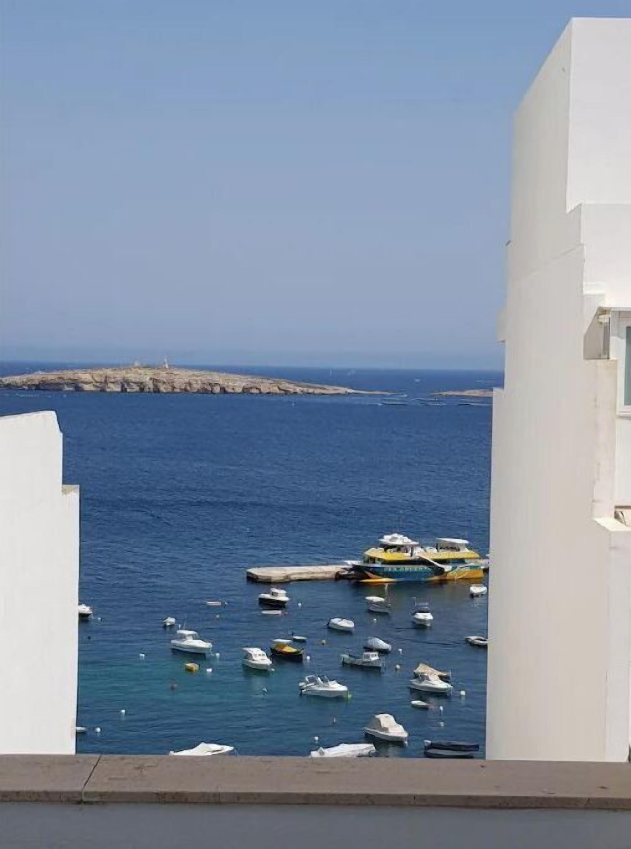 Vista costeira com barcos atracados num cais, um farol ao longe, e edifícios brancos emoldurando a paisagem.