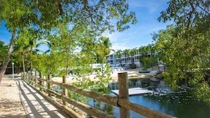 A wooden bridge over a calm water body, surrounded by lush greenery and modern buildings in the background. - Overlooking the Lagoon and Pool (Key Largo)