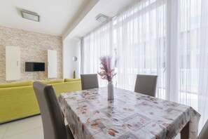 Ein modernes Wohnzimmer mit einer Couch, einem Esstisch mit floralem Tischläufer und einem großen Fenster mit durchsichtigen Vorhängen.