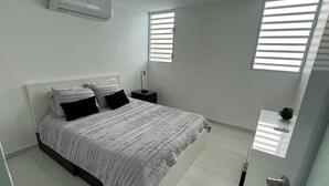 A bedroom with a bed, a nightstand, a small table with a plant, and a door. - Ocean Side PR (Carolina)