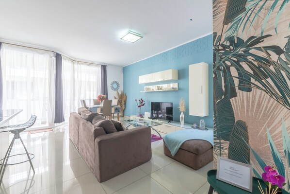 A modern living room with a sofa, a glass coffee table, and a TV. There is a dining area with a table and chairs.