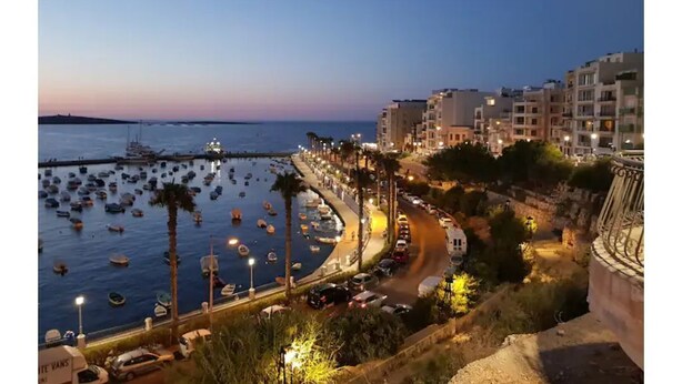 A coastal cityscape at dusk with a marina filled with boats, a curved road lined with buildings, and a prominent cliff overlooking the area. - ☆☆Amazing Apartment w/side sea view☆☆ 1 (St. Pauls Bay, Malta)