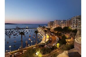 Un paisaje urbano costero al anochecer, con un puerto lleno de barcos, una carretera curva bordeada de edificios y un acantilado prominente que domina la zona.