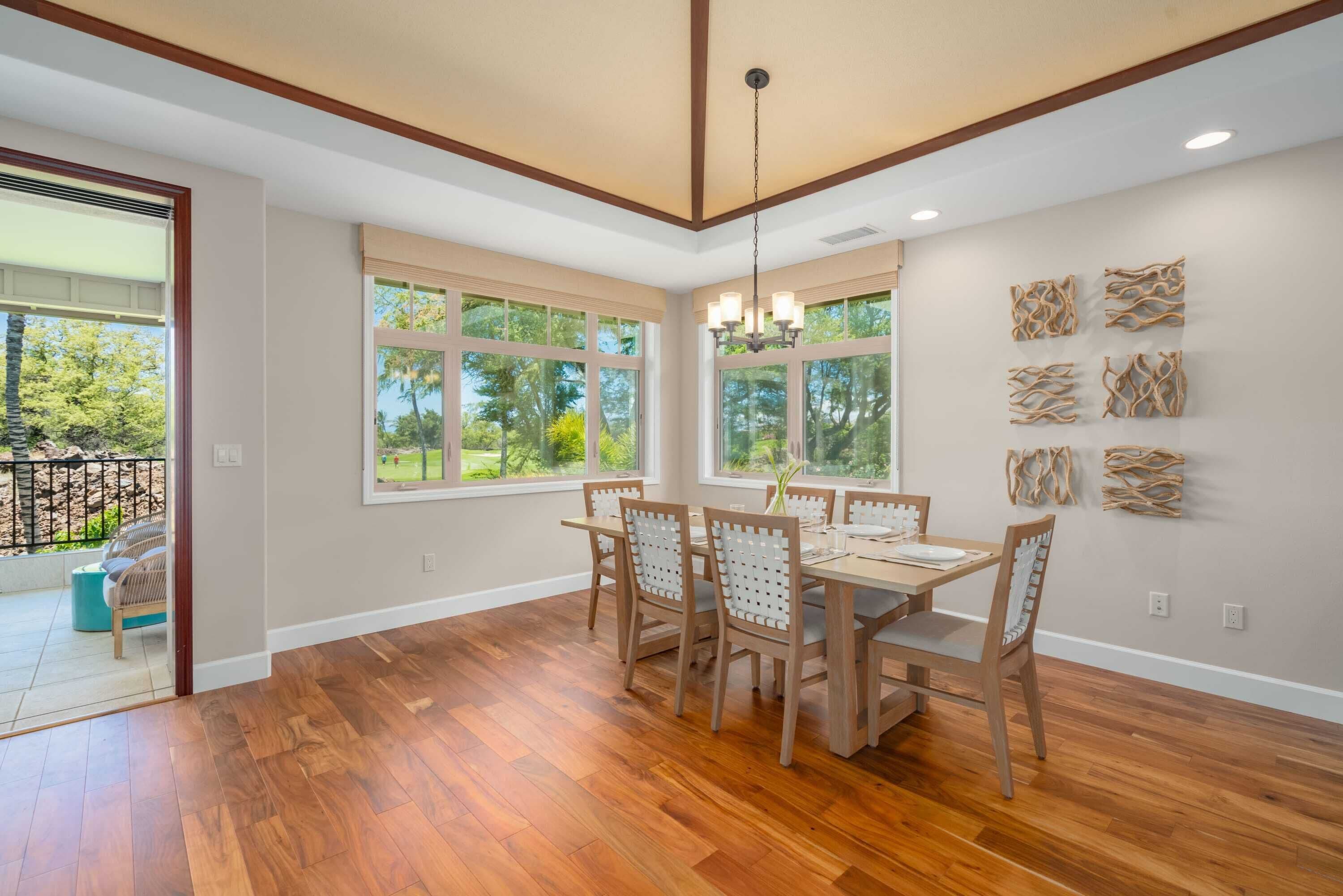 A dining area with a wooden table and chairs, large windows with a view of greenery, and a chandelier.
