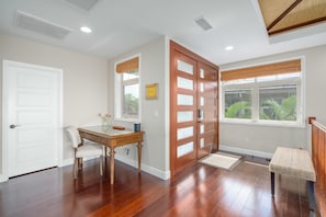 A room with hardwood flooring, a wooden desk with a chair, a white door, a glass door, and a window with a view of greenery.
