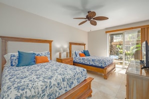 A hotel room with two beds, a ceiling fan, a TV, and a balcony with a view of greenery.