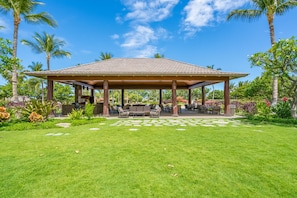 A covered outdoor seating area with a tiled roof, surrounded by lush greenery and palm trees.