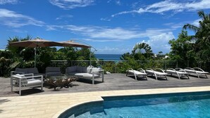 A poolside area with lounge chairs and a table under an umbrella, overlooking a body of water. - Villa Anse Vinaigri - Beach on foot 👣 (Le Gosier)