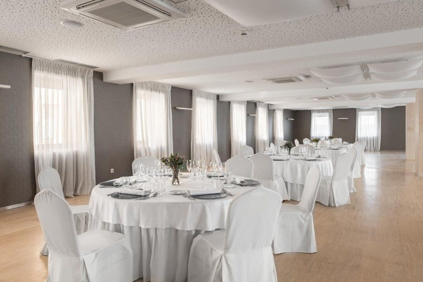 A spacious banquet hall with round tables set for dining, white chairs, and large windows with sheer curtains.