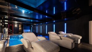 Un área de piscina interior moderna con techo iluminado en azul, una piscina y tres sillones beige para relajarse.