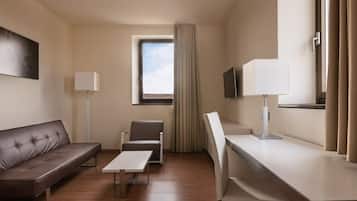 A hotel room with a brown leather sofa, a small table, a chair, a desk with a lamp, a TV, and a window with curtains.