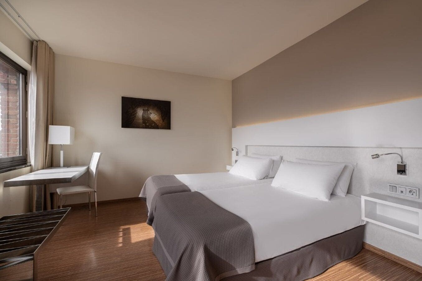 A hotel room with a bed, a desk, a chair, and a painting on the wall.