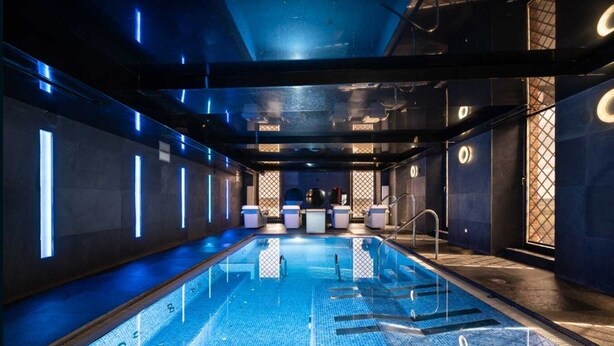 Una piscina interior moderna con un diseño largo y estrecho, rodeada de paredes con tiras de luz verticales y un área de descanso con sillones de masaje.