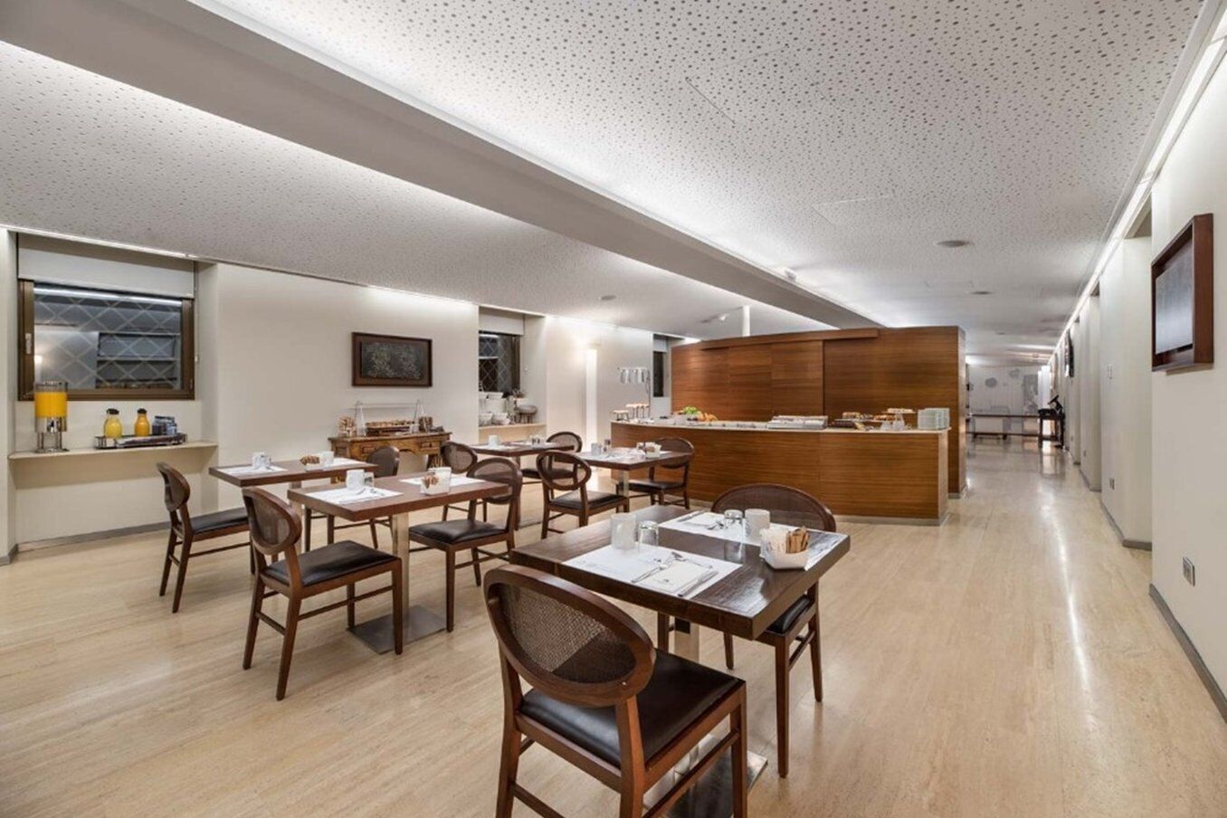 A modern hotel dining area with wooden tables and chairs, a central buffet station, and a long corridor with a polished floor.