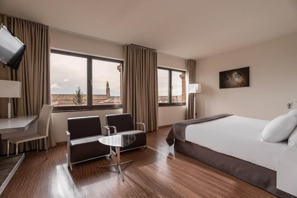 Hotelkamer met een bed, twee stoelen, een klein tafeltje, een bureau, een televisie en grote ramen met uitzicht op de stad.