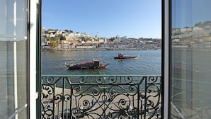 Une vue depuis un balcon donnant sur une rivière où naviguent des bateaux, avec une vue sur la ville en arrière-plan et une rambarde métallique au premier plan.
