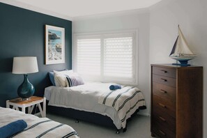 A bedroom with a bed, a dresser, a lamp, a window with blinds, and a sailboat model. - La Ronde Three - Oceanstays - Unit 3 (Yamba)