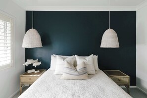 A bedroom with a dark wall, a bed with white bedding, two hanging light fixtures, and a nightstand with decorative fish. - La Ronde Three - Oceanstays - Unit 3 (Yamba)