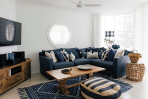 A modern living room with a sectional sofa, a wooden coffee table, a television, and a large window with blinds. - La Ronde Three - Oceanstays - Unit 3 (Yamba)