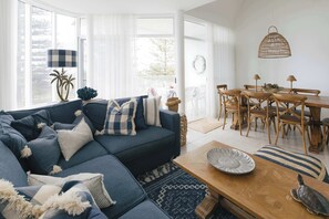 A modern living room with a sectional sofa, a wooden dining table with chairs, and a view of the outdoors through large windows. - La Ronde Three - Oceanstays - Unit 3 (Yamba)