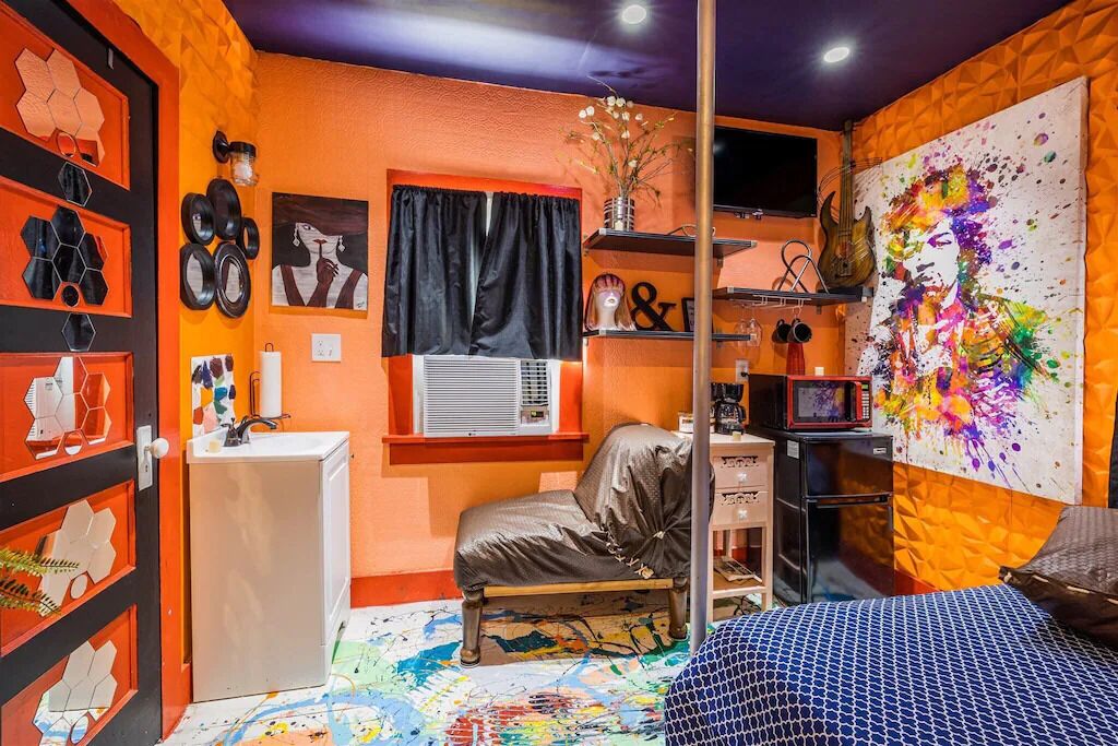 Una habitación con cama, escritorio, silla, estante con objetos decorativos y un cuadro abstracto colorido en la pared.