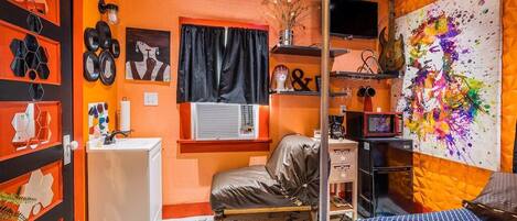 Una habitación con cama, escritorio, silla, estante con objetos decorativos y un cuadro abstracto colorido en la pared.