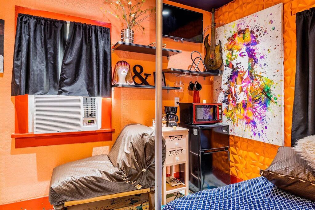 Quarto compacto com uma cama, uma pequena escrivaninha, uma prateleira com objetos decorativos e uma pintura abstrata colorida na parede.