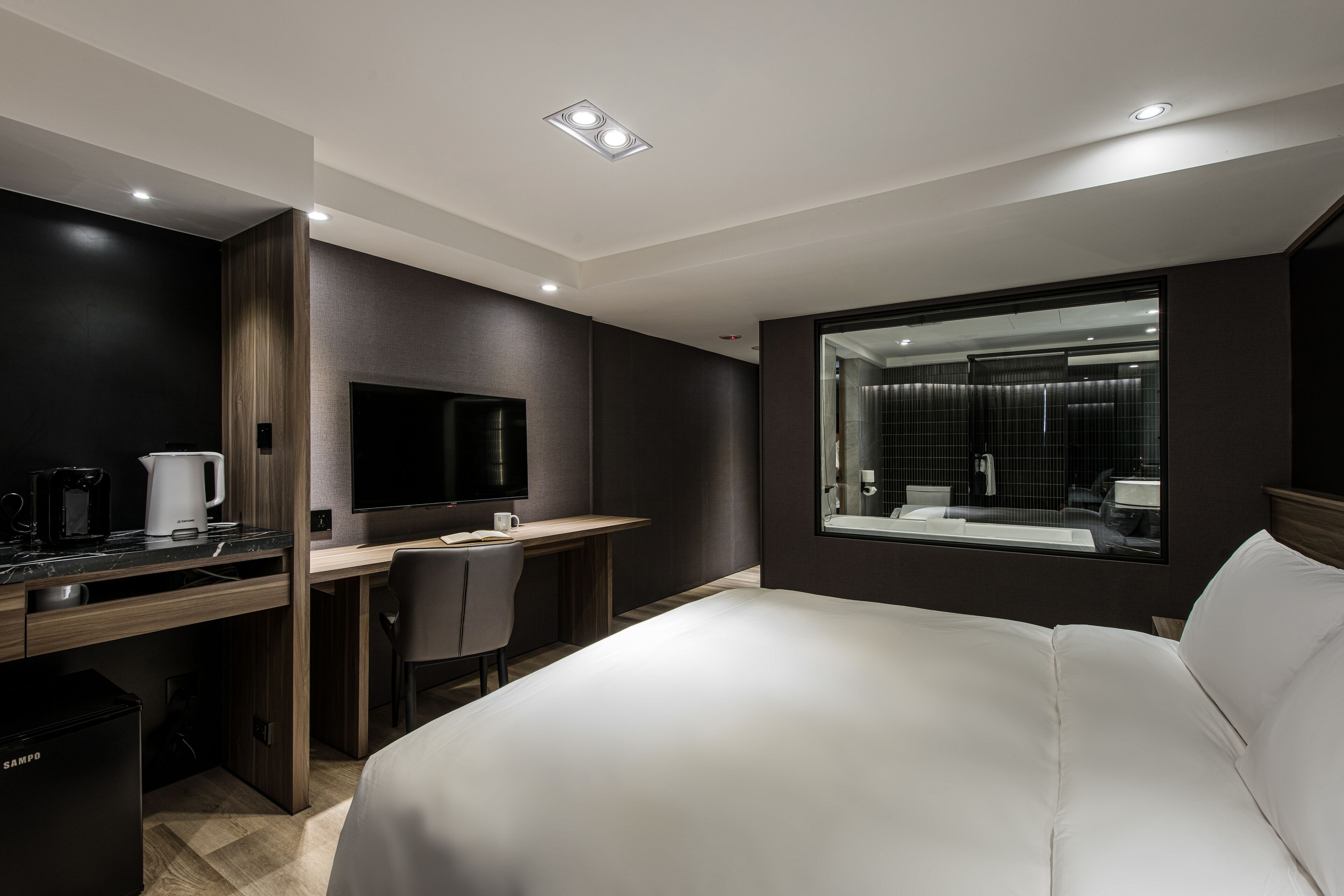 A modern hotel room with a large bed, a desk with a flat-screen TV, a coffee maker, and a view of the bathroom through a large window.