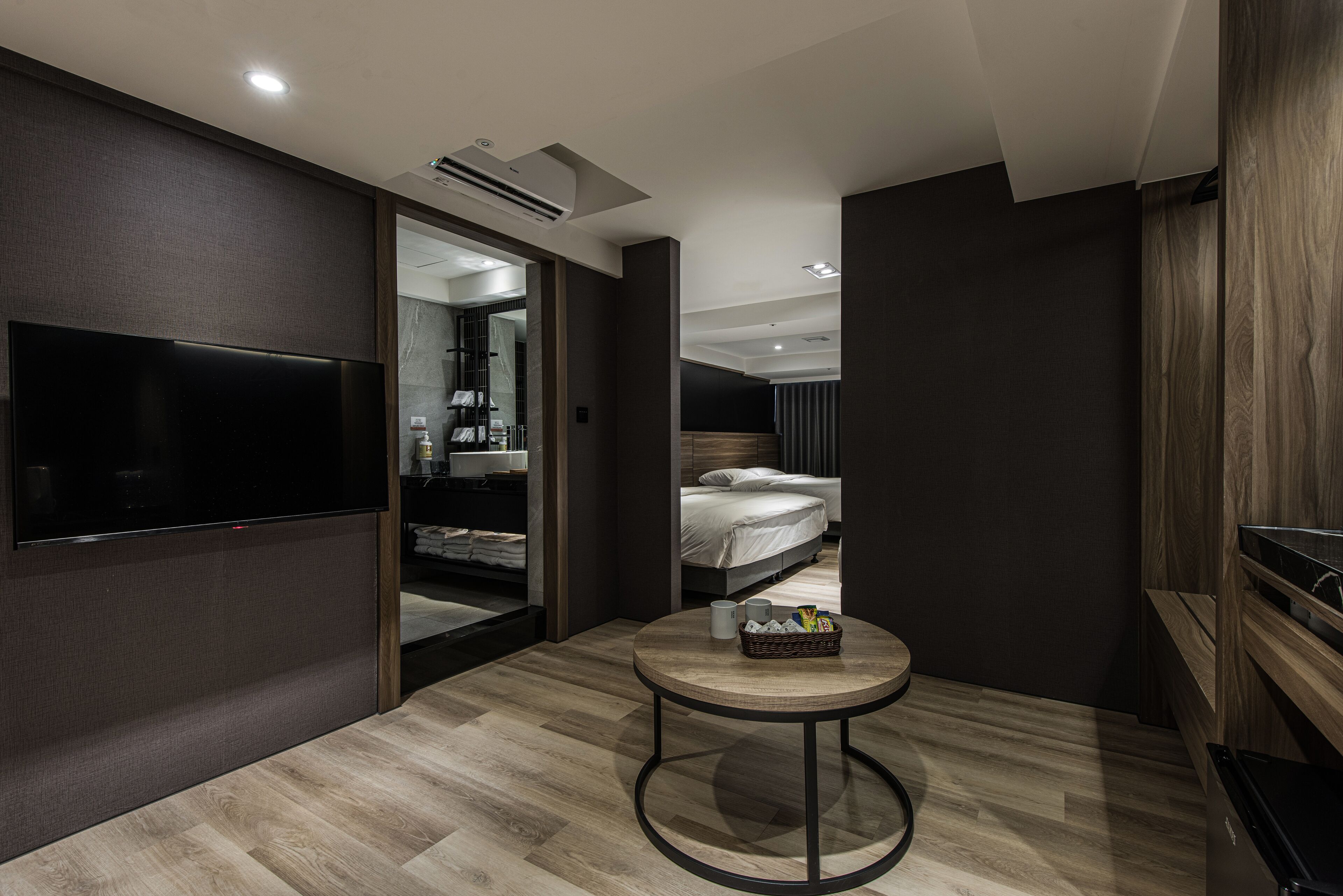 A modern hotel room with a large flat-screen TV, a round wooden table with a basket, and a bed with a headboard.