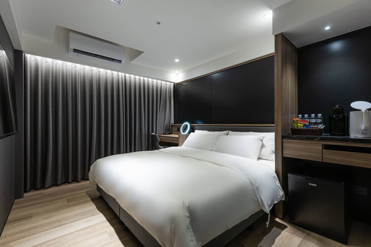 A modern hotel room with a large bed, a desk with a lamp, a mini-fridge, and a wall-mounted TV.
