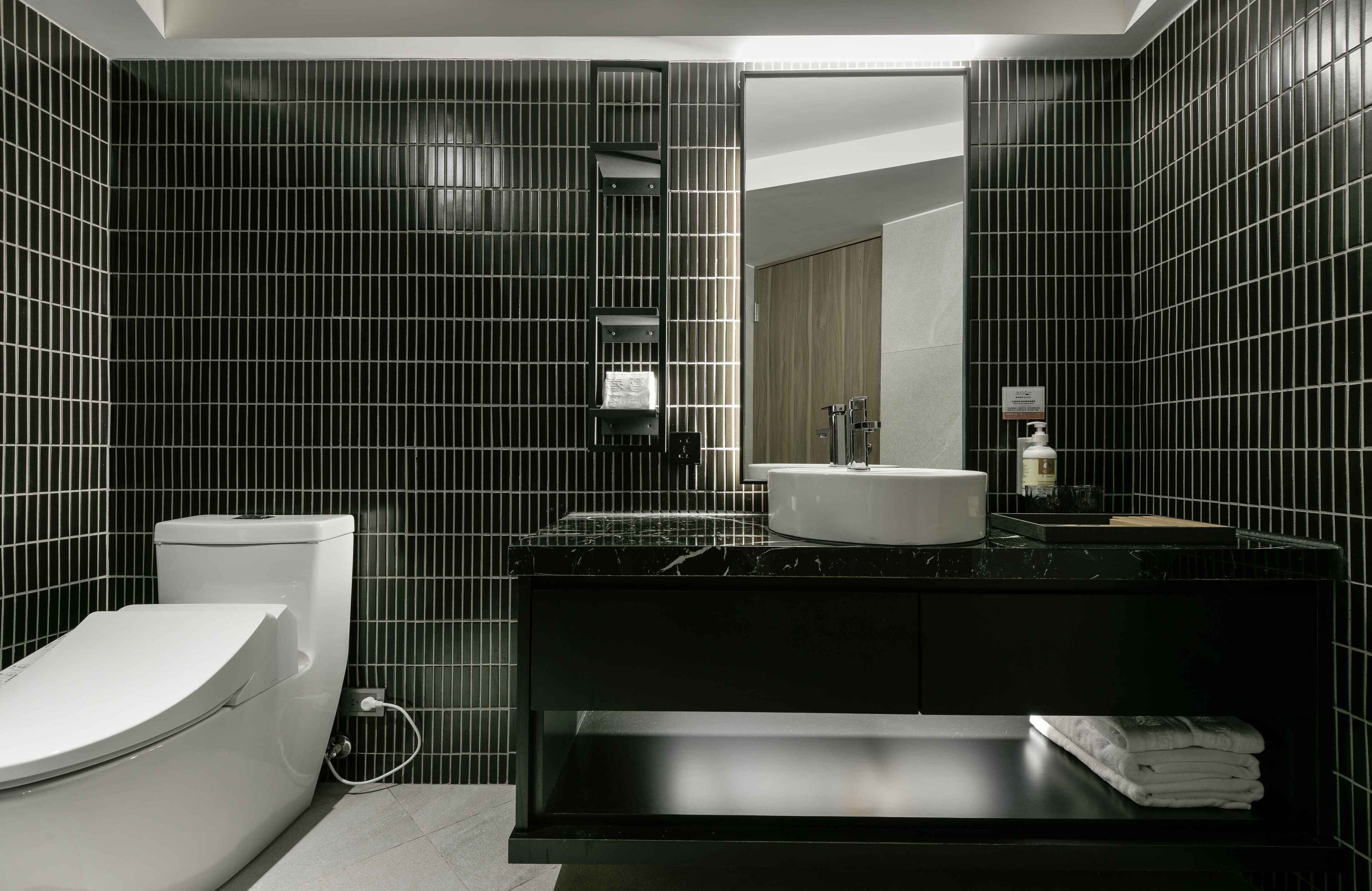 A modern bathroom with black tiled walls, a white toilet, a sink, and a mirror.