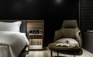 A modern hotel room with a bed, a nightstand with a phone and a small screen, a leather armchair, a round table with a book and a mug, and a window with blinds. - Autumn Willow Hotel (Taichung)