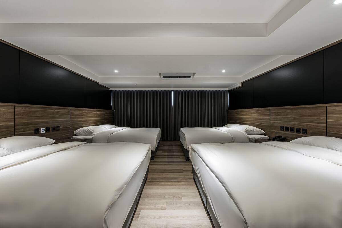 A modern hotel room with two single beds, a wooden headboard, a dark curtain, and a ceiling-mounted air conditioning unit.