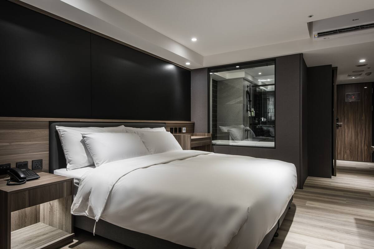 A modern hotel room with a large bed, a bedside table with a phone, and a large window offering a view of the interior.
