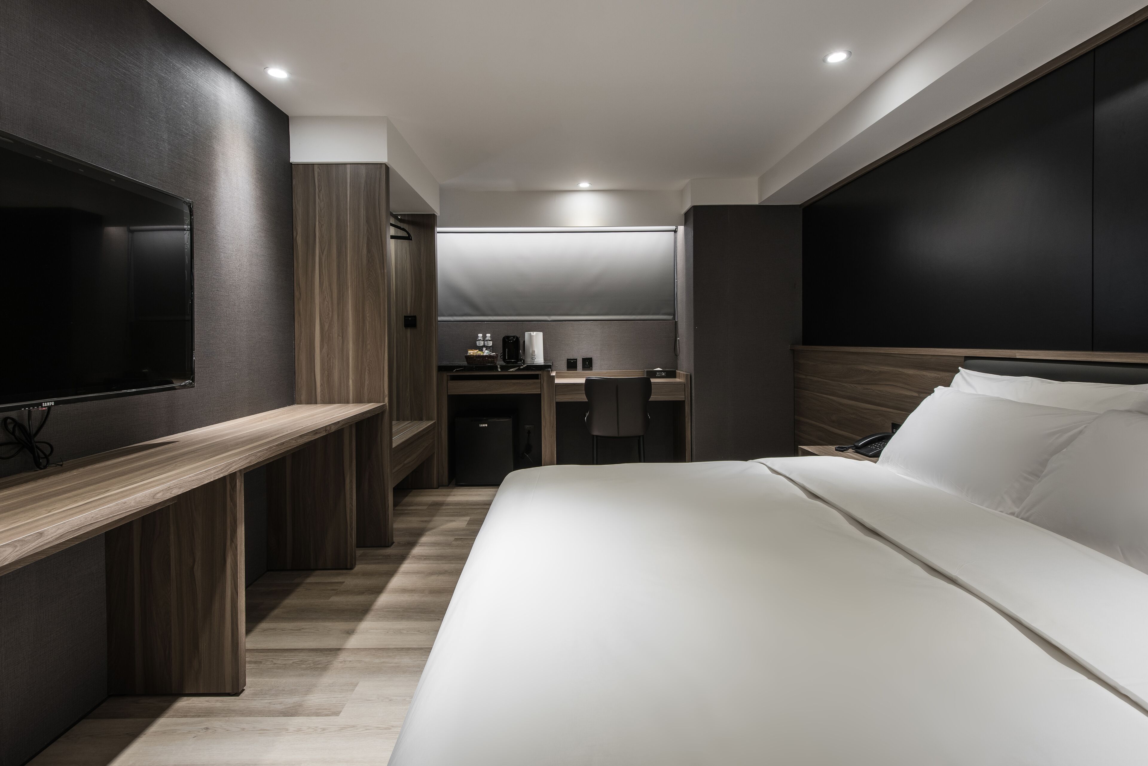 A modern hotel room with a large bed, a TV mounted on the wall, a desk, and a kitchenette.