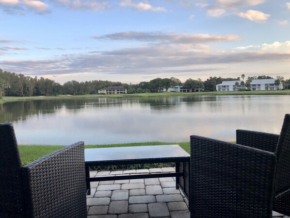 A tranquil lakeside patio with wicker furniture and a table, overlooking a calm lake and distant buildings.