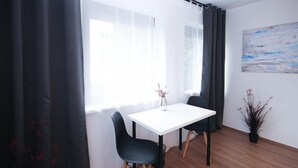 A room with a white table and two black chairs, a window with sheer curtains, a painting on the wall, and a potted plant.