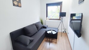 A modern living room with a dark grey sofa, a small round table with a vase, a floor lamp, and a flat-screen TV mounted on the wall.