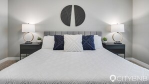 Ein ordentlich bezogenes Bett mit weißer Daunendecke, zwei Nachttische mit Lampen und eine rundes Wandbild.