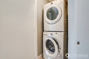 Pyykinpesuhuoneessa on päällekkäin asennettu pesukone ja kuivausrumpu. Seinät ovat valkoiset ja lattia on laatoitettu.