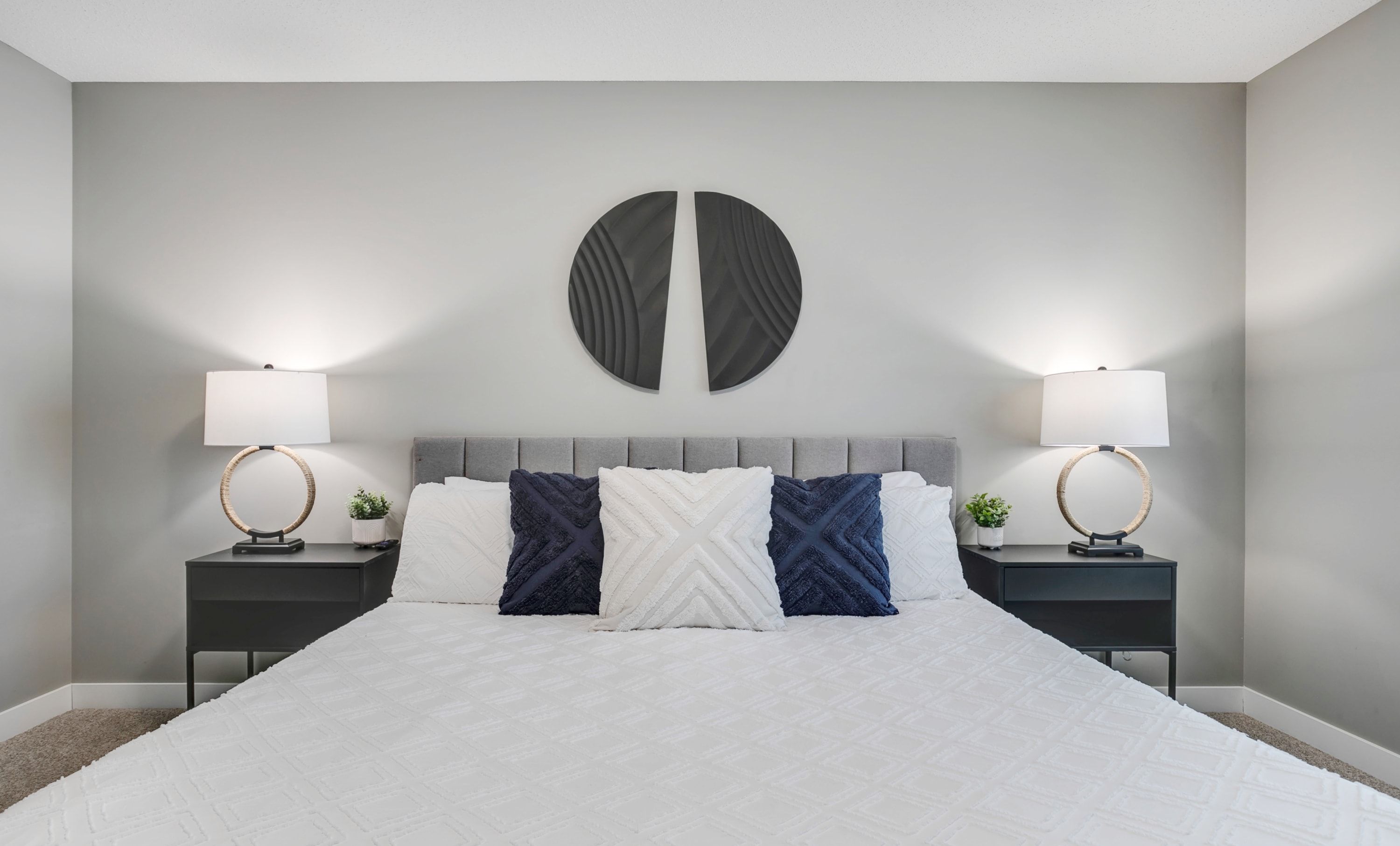 A neatly made bed with a white duvet, two bedside tables with lamps, and a wall-mounted circular decoration.