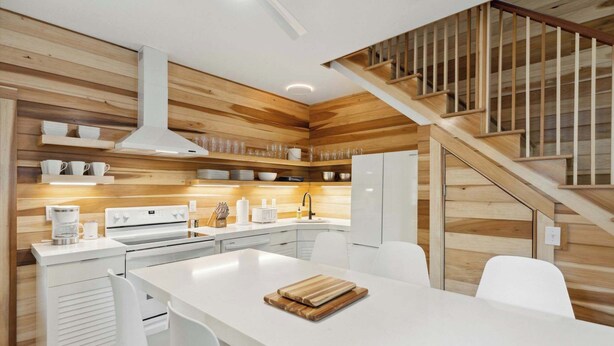 A modern kitchen with wooden paneling, white cabinetry, a central island, and a staircase with wooden railings. - ‘Sand Swept’ - Downtown Private Cottage (Charleston)