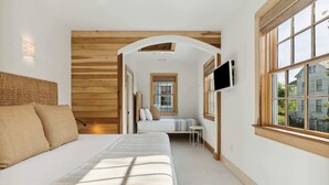 A bedroom with a large bed, a TV mounted on the wall, and a window with a view of a house. - ‘Sand Swept’ - Downtown Private Cottage (Charleston)