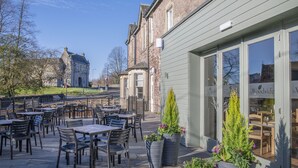 Een terras buiten met tafels en stoelen, een stenen gebouw op de achtergrond en een restaurant met een bord.