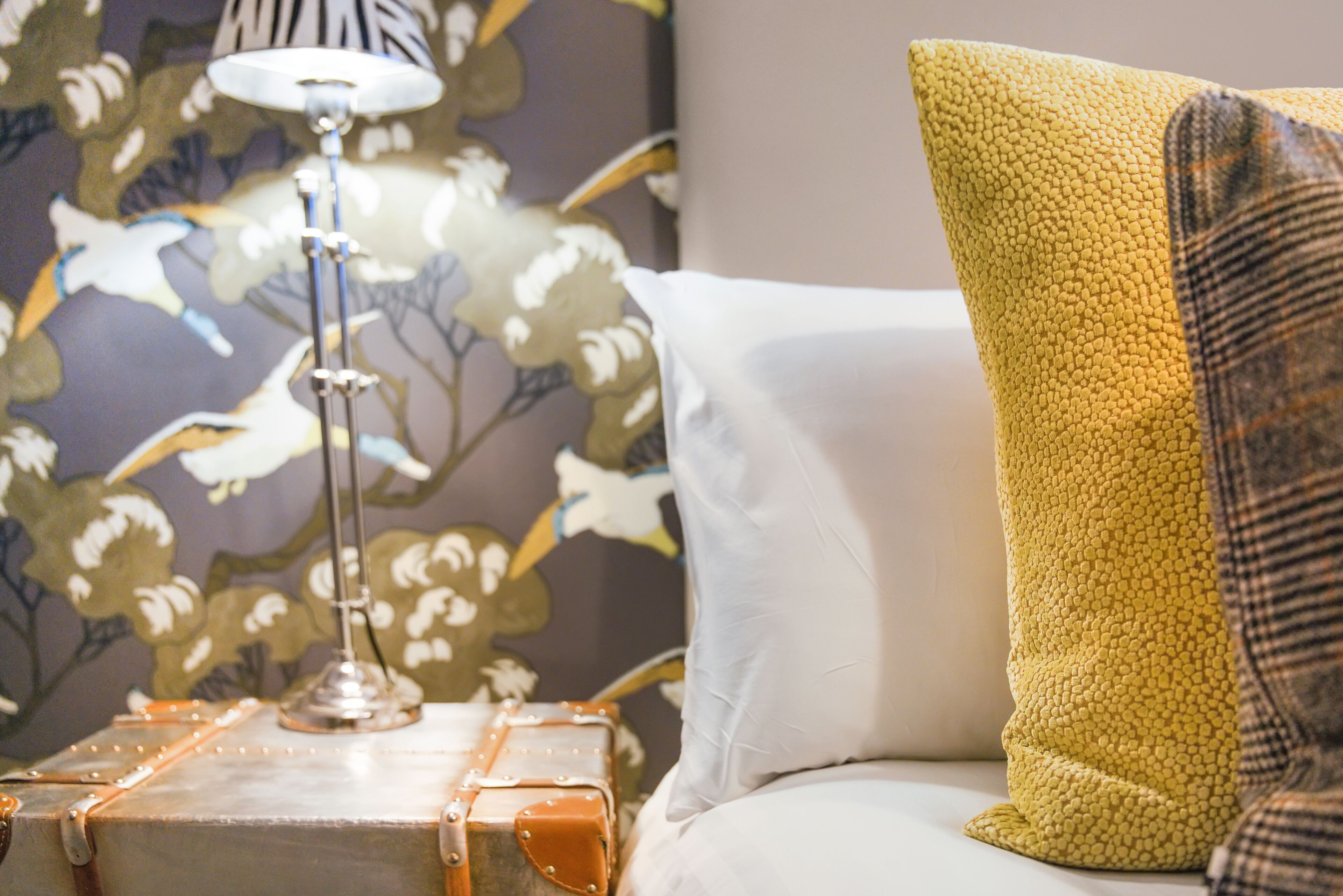 Una cama con un almohadón blanco y un almohadón amarillo texturizado, una lámpara sobre una mesita de noche, y un papel tapiz floral al fondo.