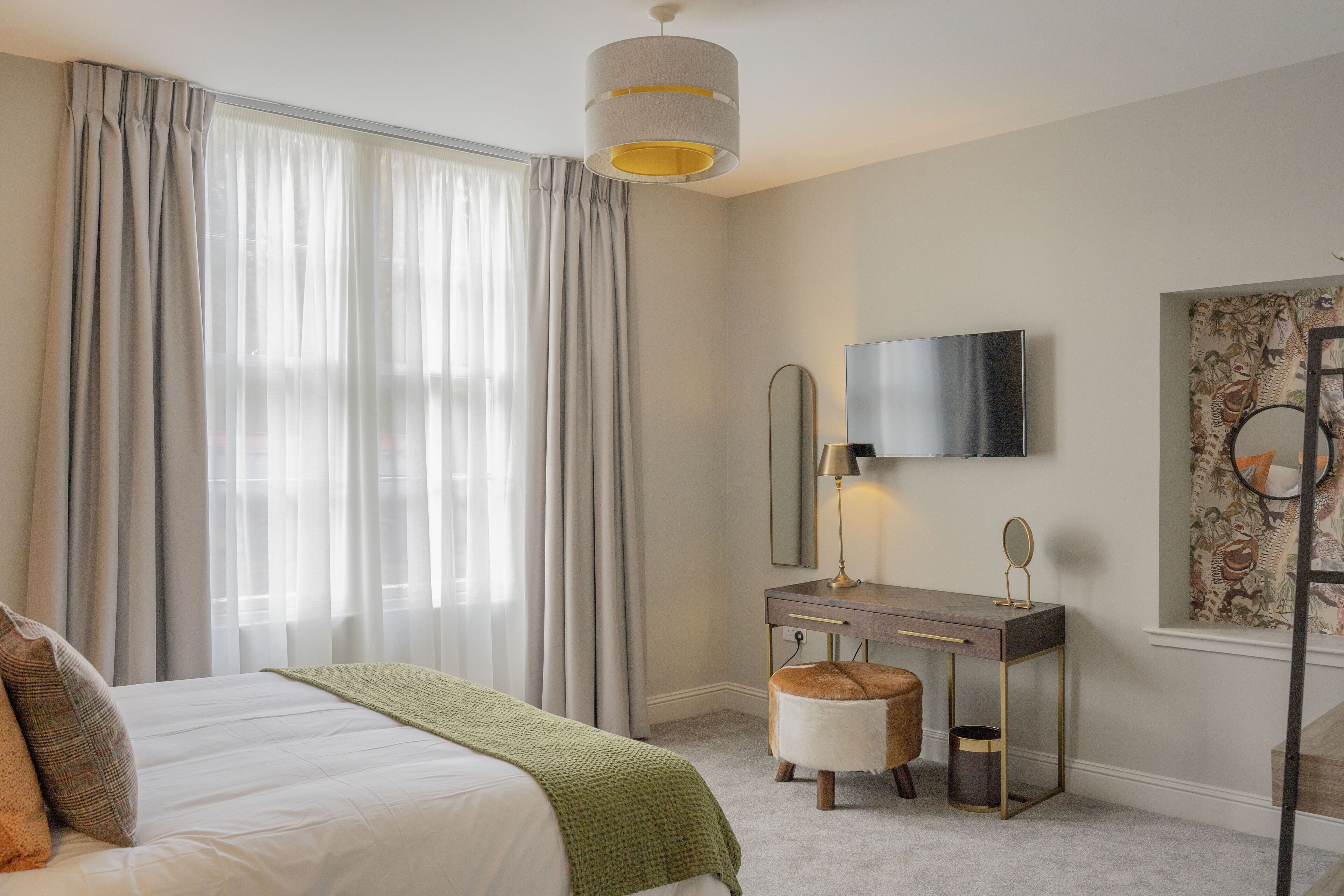 Quarto com cama, penteadeira com espelho, luminária e televisão na parede.