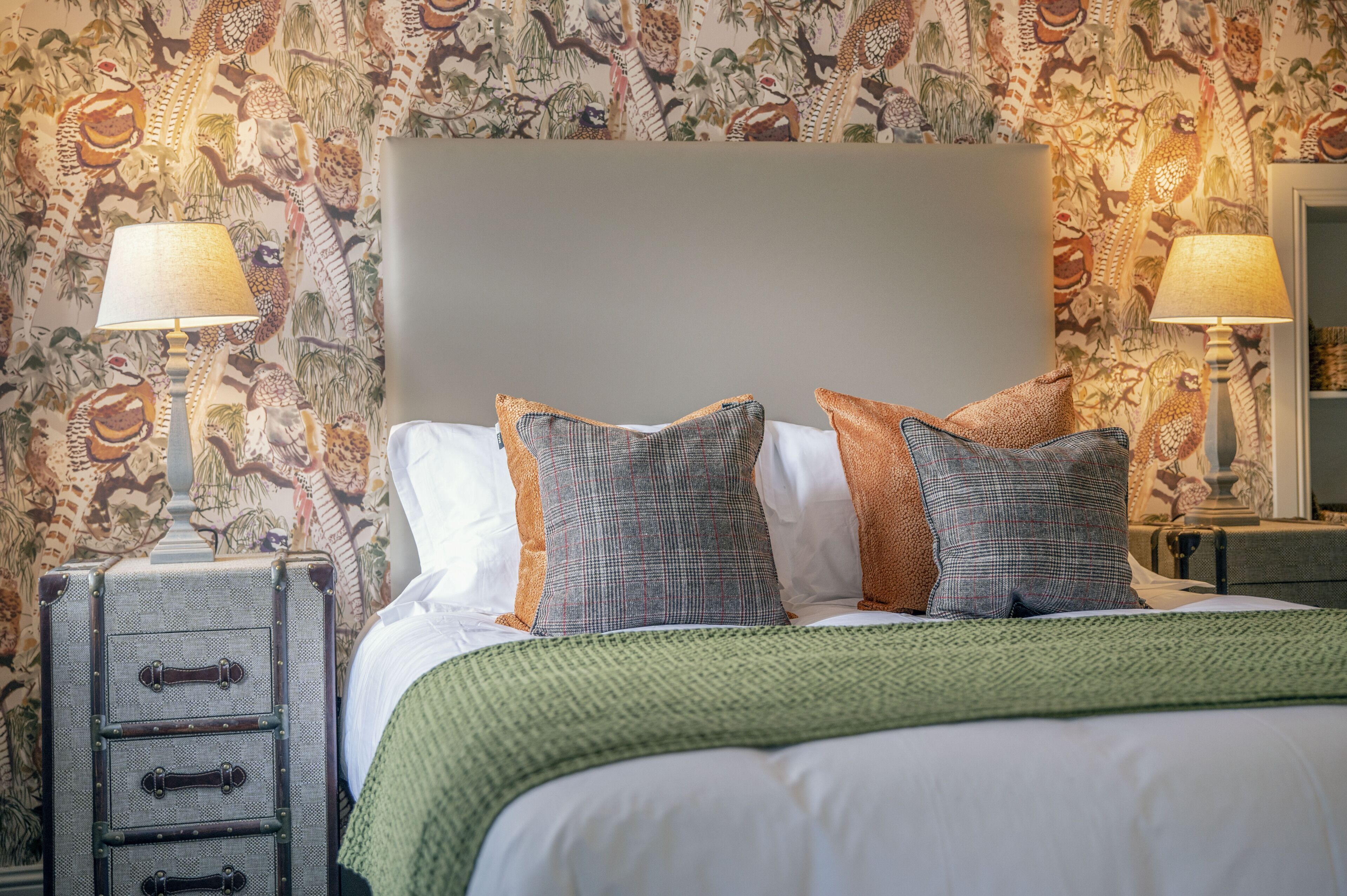 A neatly made bed with a green blanket, two lamps on nightstands, and a patterned wallpaper.
