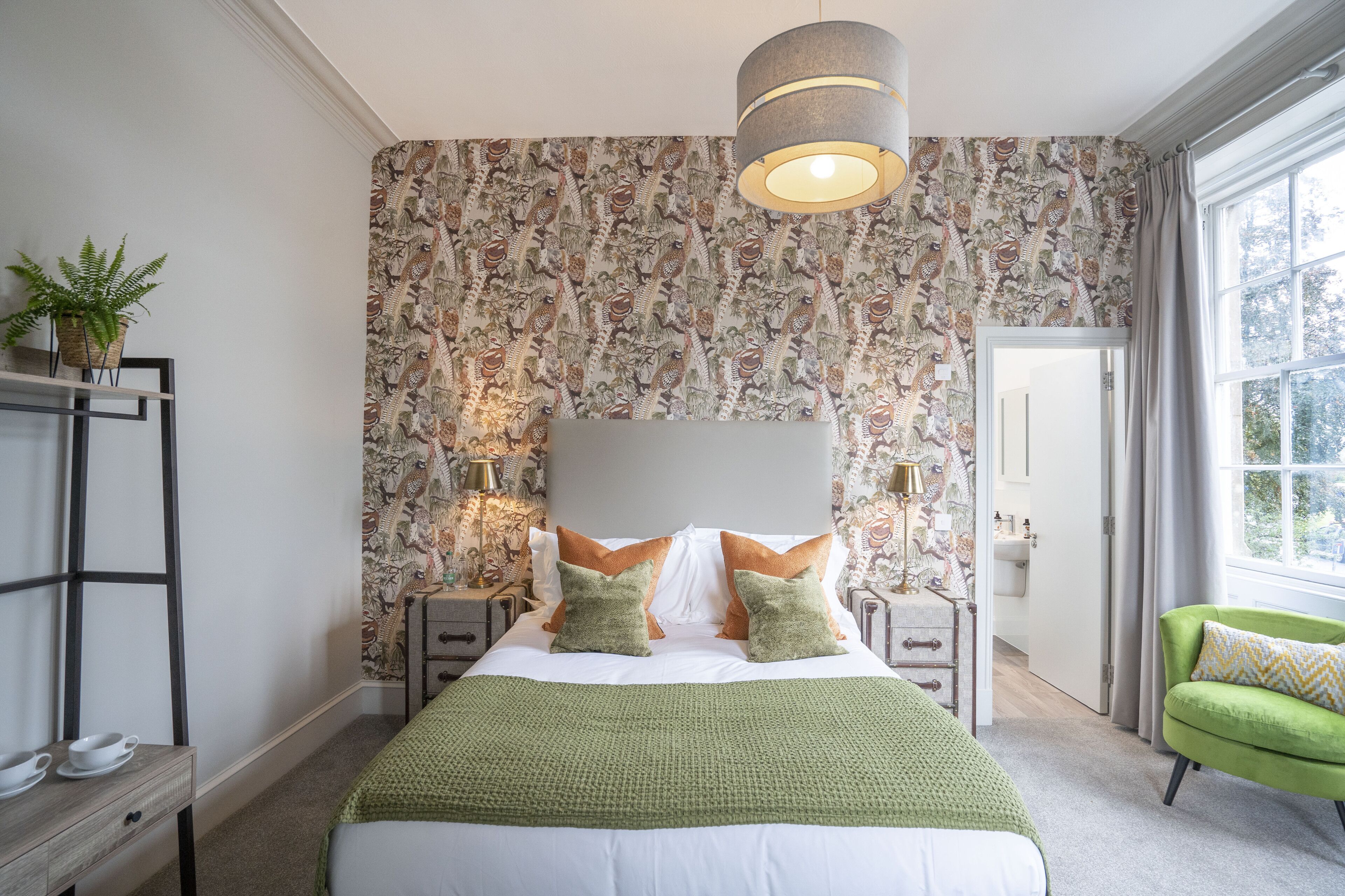A bedroom with a large bed, green throw, patterned wallpaper, bedside lamps, and a view of the outdoors.