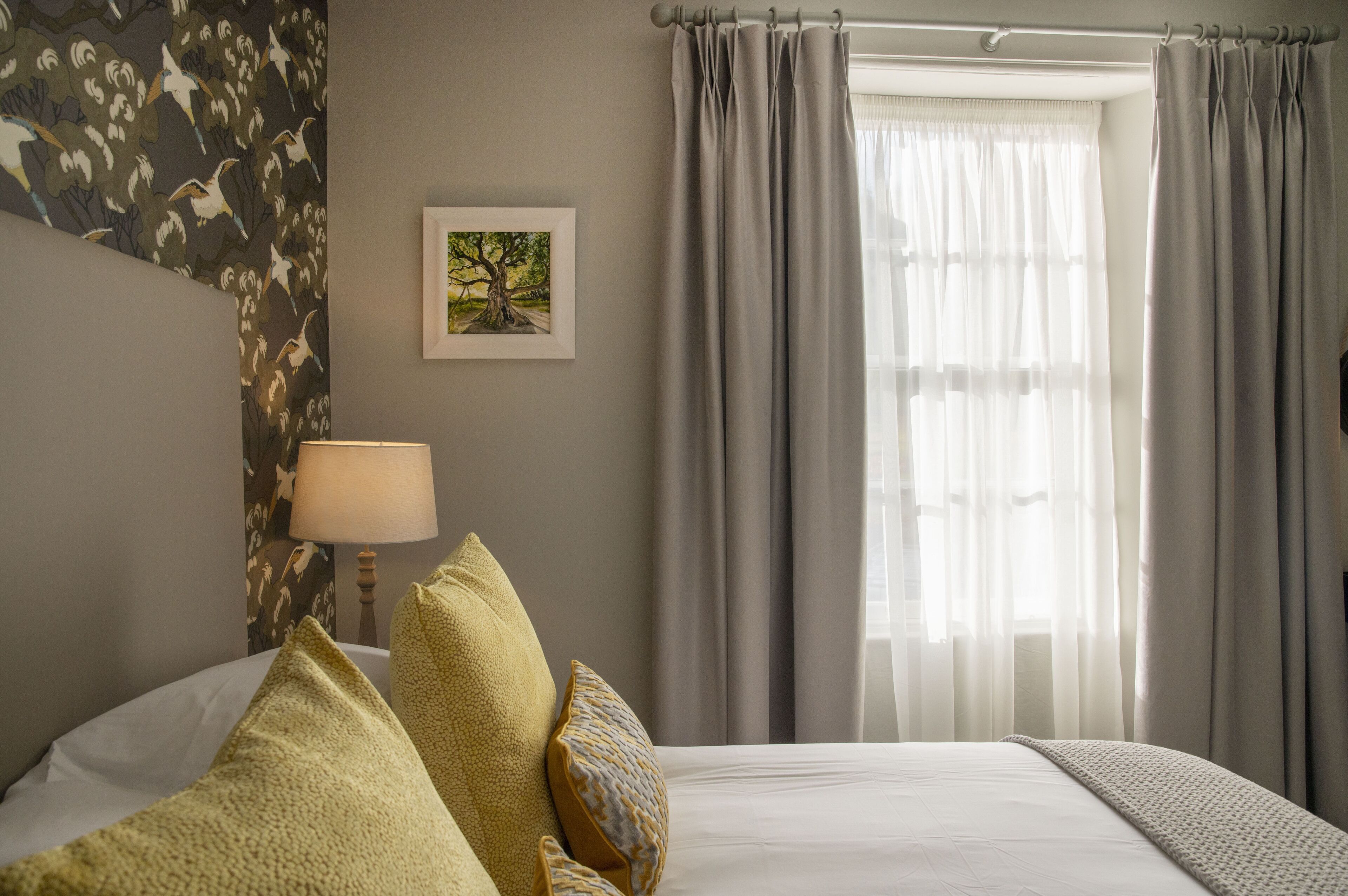 A bedroom with a bed, pillows, a lamp, a framed picture, and curtains.